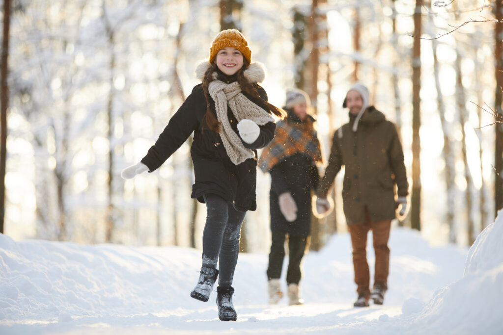 Happy little girl running towards camera in winter forest while enjoying walk with family